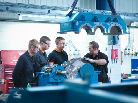 Instructor showing three apprentices a machine component in a training workshop